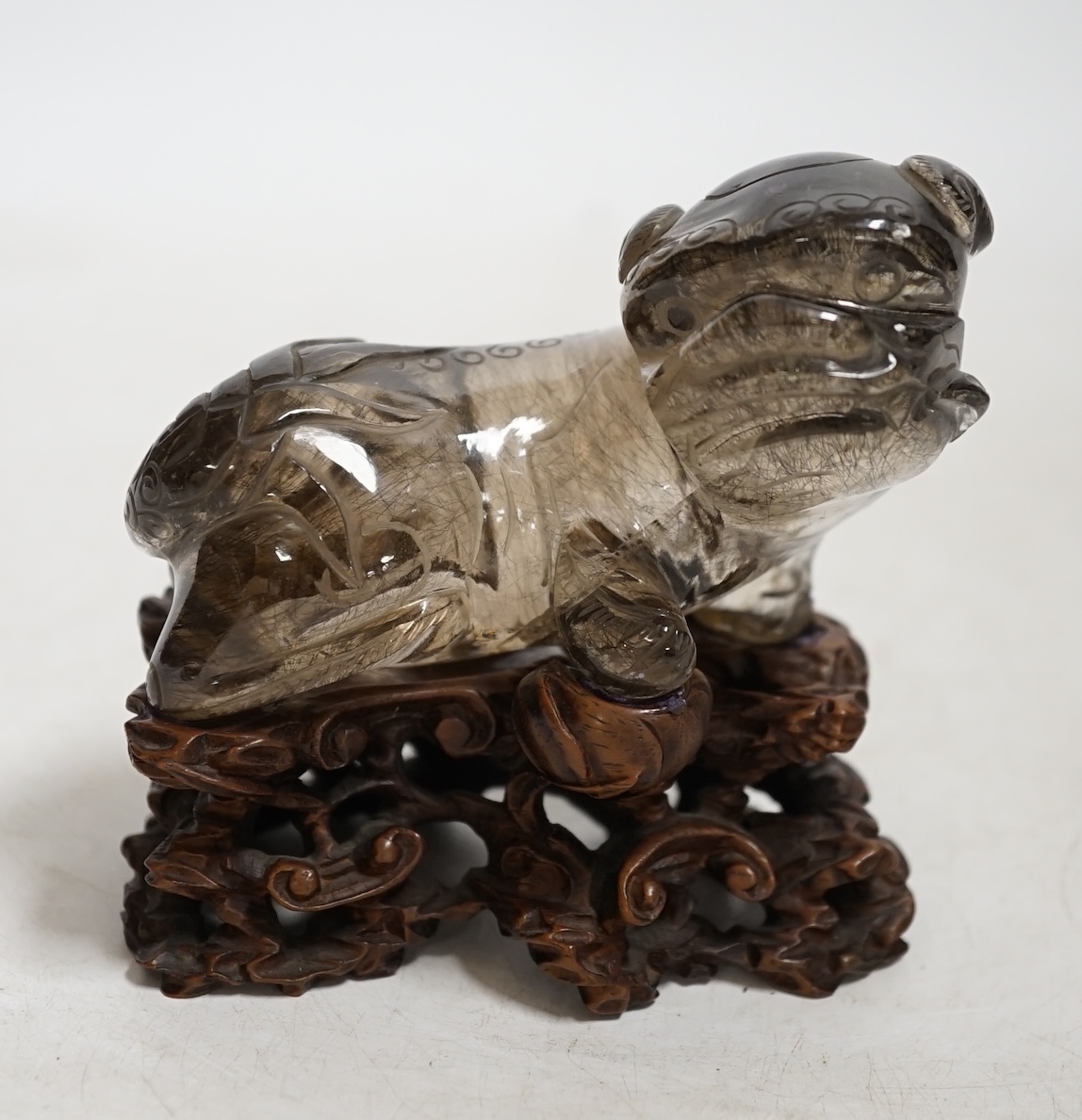 A Chinese carved smoky quartz lion dog on hardwood stand, 9.5cm wide. Condition - fair, impact damage to tail